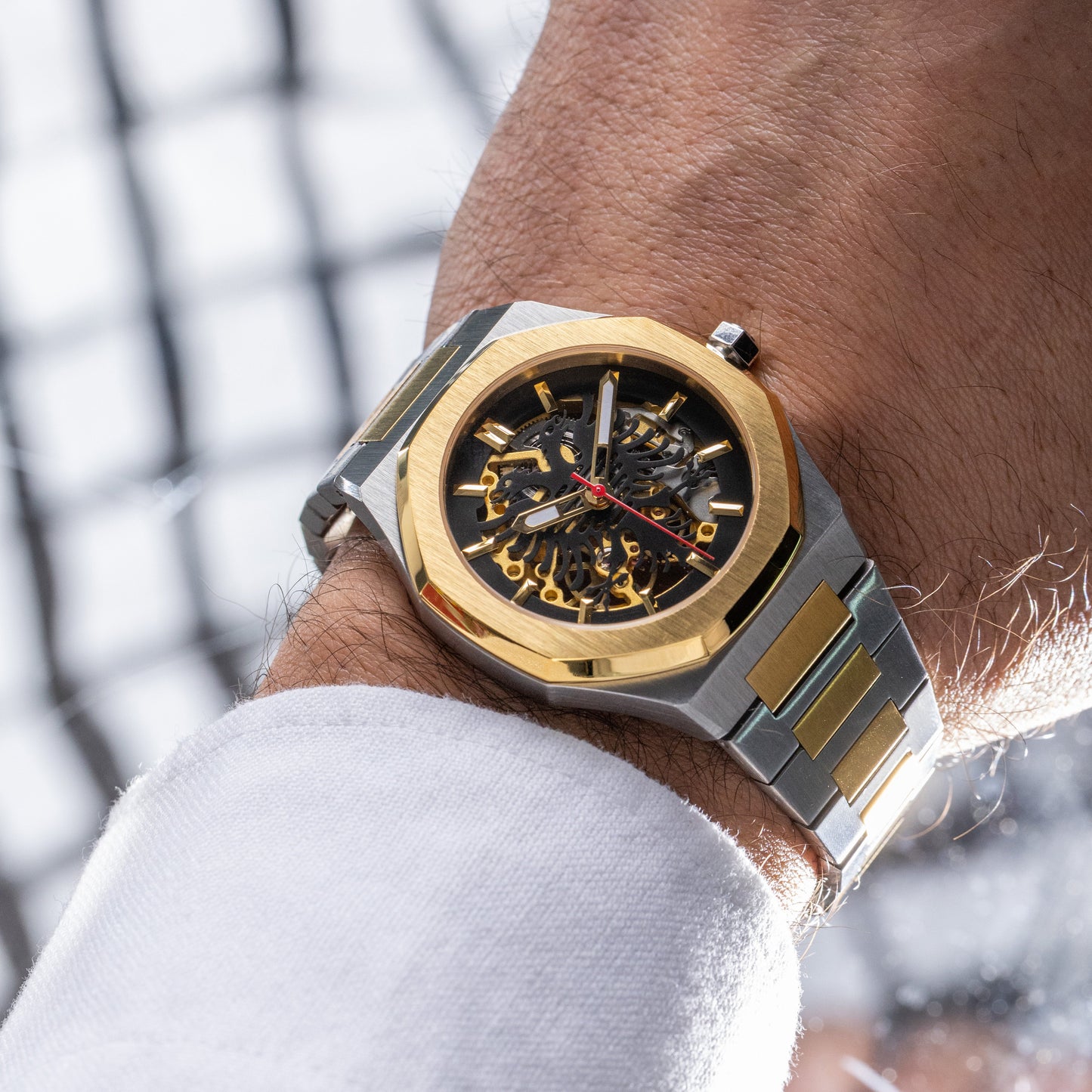 Close-up of a wrist wearing a gold and silver Albanian Eagle watch with a blurred background